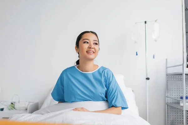 joyful asian woman in hospital gown sitting on bed in clinic and looking away