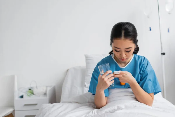 病院のベッドでコップ一杯の水を飲みながら丸薬容器を見ている若いアジア人女性 — ストック写真