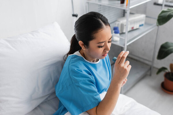 high angle view of sick asian woman holding inhaler while sitting on bed in hospital ward
