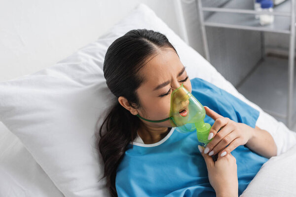high angle view of worried asian woman with closed eyes breathing in oxygen mask on hospital bed