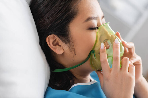side view of diseased asian woman adjusting oxygen mask in hospital ward