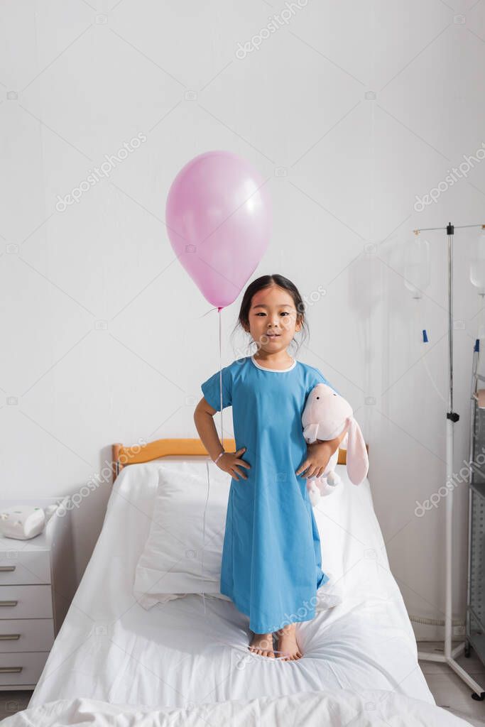 Full length of asian girl in hospital gown standing with toy bunny and festive balloon on bed in pediatric clinic