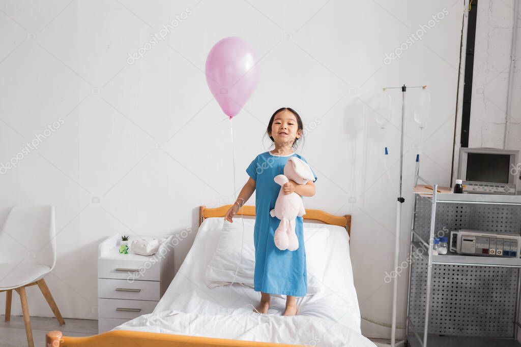 Full length of cheerful asian child standing on hospital bed with toy bunny and festive balloon