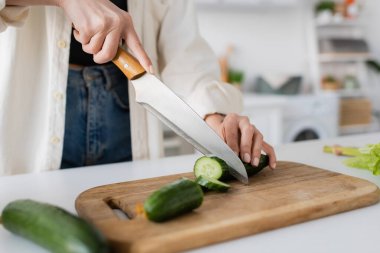 Cropped view of woman cutting ripe cucumber on chopping board in kitchen  clipart