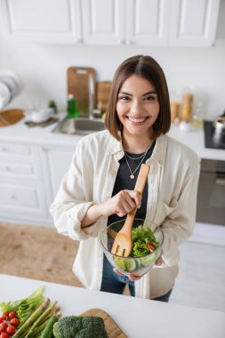 Pozitif genç kadın taze salata tutuyor ve mutfaktaki kameraya bakıyor. 