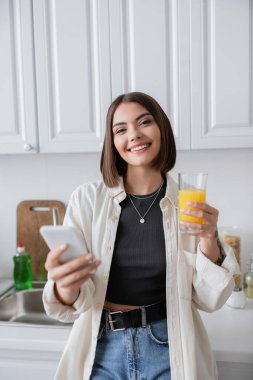 Mutfakta kameraya bakarken elinde portakal suyu ve akıllı telefon tutan gülümseyen genç bir kadın. 