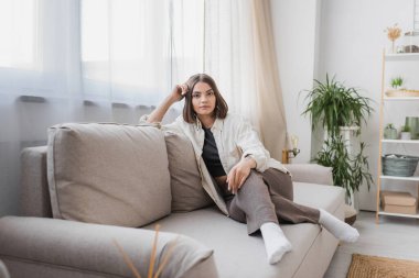 Brunette woman in casual clothes looking at camera while sitting on couch at home  clipart