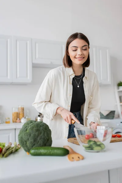 台所でサラダを調理しながら野菜を切る若い女性の笑顔 — ストック写真