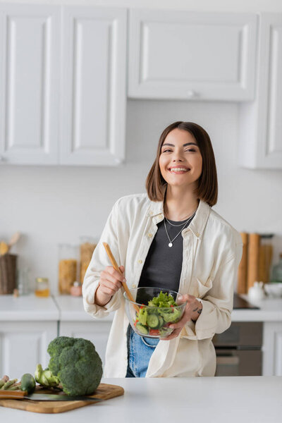 Positive woman mixing fresh and vegetarian salad in kitchen 