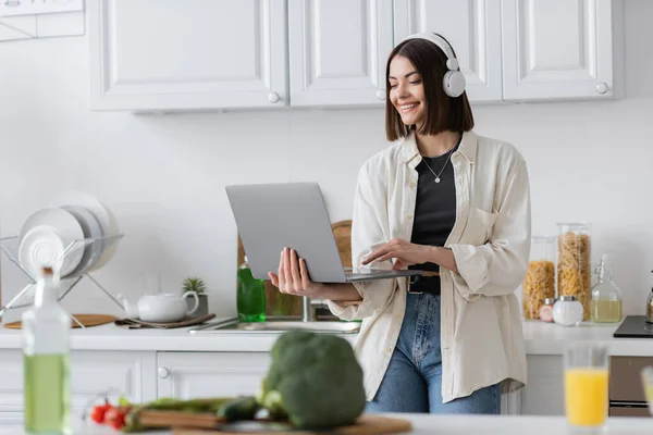 无忧无虑的女人戴着耳机 在厨房里靠近模糊的蔬菜和橄榄油的地方使用笔记本电脑 — 图库照片