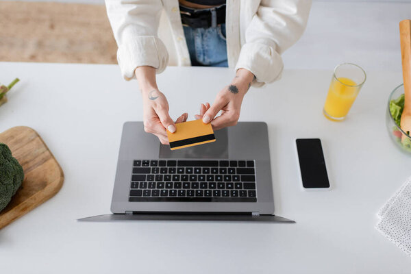 Вид сверху татуированной женщины, держащей кредитную карту рядом с устройствами и салатом на кухне 