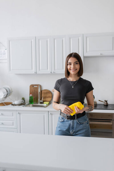 Smiling tattooed woman holding rag and looking at camera in kitchen 