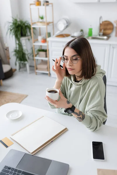 Tattooed woman in eyeglasses holding coffee cup near devices and credit card on table at home