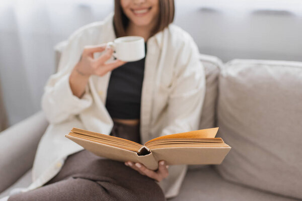 Cropped view of blurred woman holding coffee cup and reading book on couch at home 
