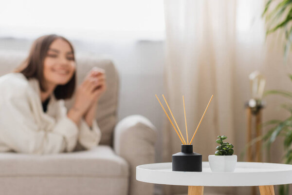 Plant and bamboo scented sticks on coffee table near blurred woman on couch at home 