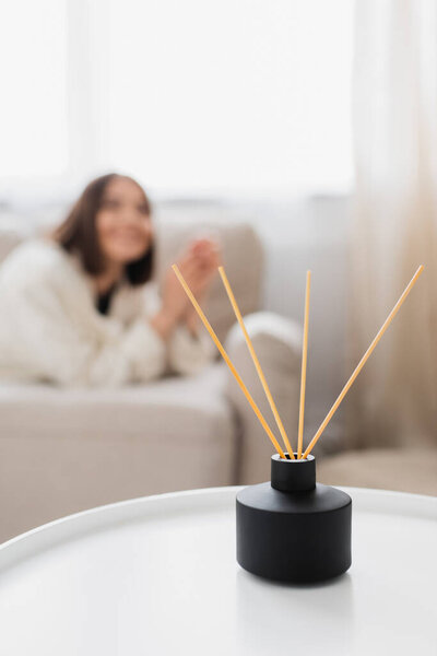 Aroma diffuser with bamboo sticks on coffee table near blurred woman in living room