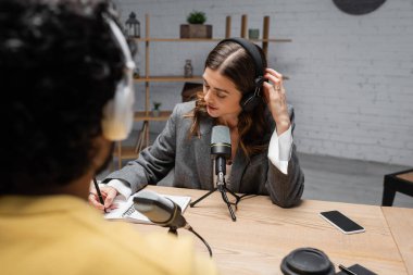 charming broadcaster in headphones and grey blazer writing in notebook near microphones, smartphone with blank screen and paper cup during interview with blurred indian man in radio studio clipart