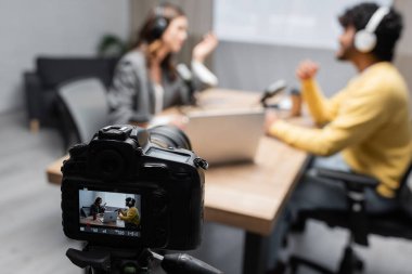 selective focus of professional digital camera recording how interviewer talking to young indian guest while sitting together near laptop and microphones on blurred background in studio clipart