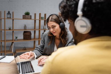 smiling brunette radio host in headphones and grey blazer pointing at laptop near notebook and young indian guest in yellow jumper on blurred foreground clipart