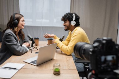 Kıvırcık saçlı, kulaklıklı, sırıtan Hintli ve sarı kazaklı bir adamın laptopun, defterin ve radyo stüdyosundaki bulanık dijital kameranın önünde esmer bir röportajcıyla konuşmasının yan görüntüsü.