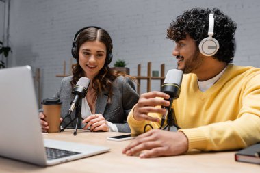 Kulaklıklarda, bilgisayarın yanında profesyonel mikrofonlarla çalışan ırklar arası yayıncının gülümsemesi, radyo stüdyosunda podcast yaparken cep telefonuyla boş ekran ile kahve içmek.