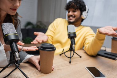 radio host pointing at takeaway drink near studio microphones, smartphone with blank screen and young indian colleague in yellow jumper smiling and showing shrug gesture on blurred background clipart