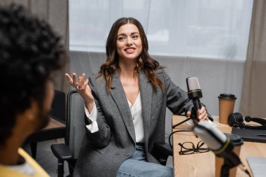 Gri ceketli, elinde mikrofon, not defteri, kağıt bardak ve gözlük tutan kumral podcaster stüdyodaki bulanık ön planda Hintli bir adamla konuşurken,