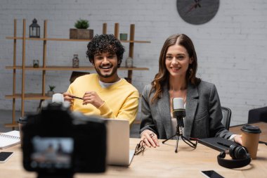 positive interracial podcasters in yellow jumper and grey blazer smiling near laptop, microphones, headphones, coffee to go and blurred digital camera in professional broadcasting studio clipart