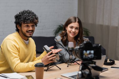 joyful and curly indian man in yellow jumper talking in microphone and pointing at charming colleague near laptop, coffee to go and smartphones in front of blurred digital camera in broadcasting studio clipart