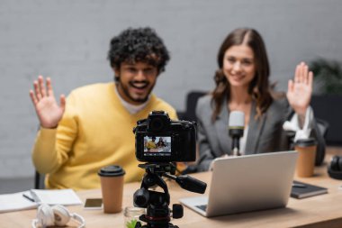 focus on digital camera in front of cheerful interracial podcasters waving hands near laptop, takeaway drinks, notebooks, headphones and mobile phone during video call in radio studio clipart