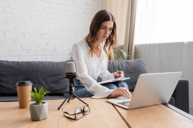 brunette woman in white blouse using laptop and writing in notebook near microphone, coffee to go, flowerpot and eyeglasses on table in professional radio studio clipart