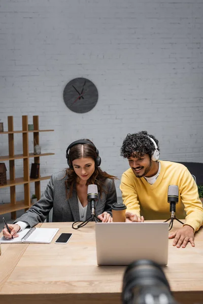 stock image smiling podcaster in headphones and blazer writing in notebook and looking at laptop near indian colleague in yellow jumper, microphones, coffee to go and smartphone in broadcasting studio