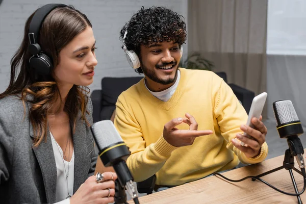 Uśmiechnięty Indyjski Podcaster Słuchawkach Żółty Sweter Pokazujący Telefon Komórkowy Uroczej — Zdjęcie stockowe
