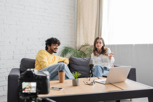 брюнетка женщина указывая на ноутбук рядом веселый индийский мужчина, стол с ноутбуком, микрофон, цветочный горшок, кофе, чтобы пойти и очки во время записи подкаста на размытой цифровой камеры в студии