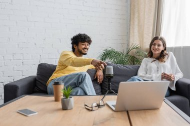 cheerful indian podcaster in yellow jumper pointing at laptop during video call near smiling colleague, microphone, coffee to go, flowerpot, smartphone and eyeglasses on table in studio clipart