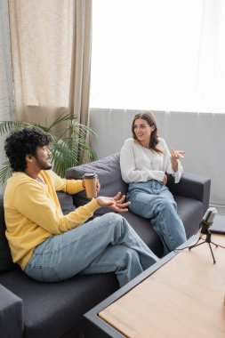 high angle view of cheerful indian man holding paper cup and talking to woman in white blouse while sitting on sofa near notebook and microphone in podcast studio clipart