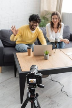 blurred indian man waving hand during video chat on laptop near smiling colleague and professional digital camera in broadcasting radio studio with paper cup and flowerpot on table clipart