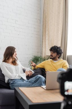 young and curly indian man in yellow jumper gesturing and talking to charming brunette woman in white blouse white sitting on couch near laptop while recording podcast in studio clipart