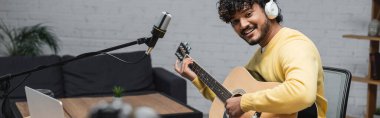 Kıvırcık ve mutlu kulaklıklı Hintli müzisyen ve sarı kazaklı akustik gitar çalan laptop ve profesyonel mikrofon stüdyoda arka planda kanepe, afiş
