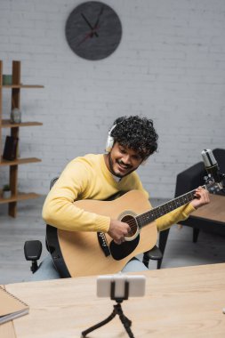 Stüdyoda podcast yaparken tripod ve profesyonel mikrofonda bulanık cep telefonunun yanında akustik gitar çalan kulaklıklı neşeli Hintli müzisyen ve sarı kazaklı.