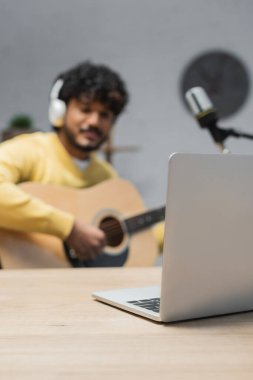 Kablosuz kulaklıklı genç Hintli adamın yanındaki tahta masaya odaklanın. Radyo stüdyosundaki mikrofonun yanında podcast yaparken akustik gitar çalın.