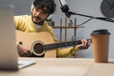 Kulaklıklı neşeli Hint podcaster 'ı ve mikrofonun yanında akustik gitar çalan sarı Jumper ve kayıt stüdyosundaki kağıt bardakta kahve içmek için bulanık dizüstü bilgisayar.