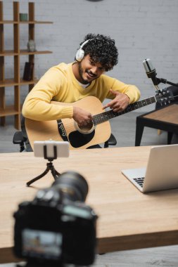 Profesyonel stüdyodaki bulanık dijital kameranın önünde dizüstü bilgisayarın ve akıllı telefonun yanında otururken akustik gitara işaret eden kulaklıklı Hintli müzisyen.