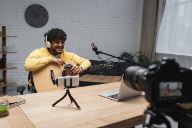 Podcast stüdyosundaki yayın sırasında mikrofon ve cihazların yanına gitmek için akustik gitar ve kahve taşıyan gülümseyen Hintli podcaster. 