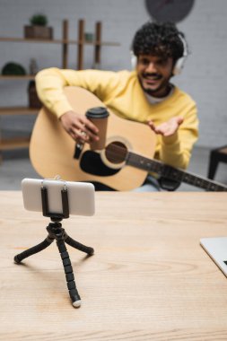 Üç ayaklı akıllı telefon, bulanık Hindistan podcaster 'ının yanında. Kulaklıklarda kahve var. Podcast stüdyosunda akustik gitar var. 