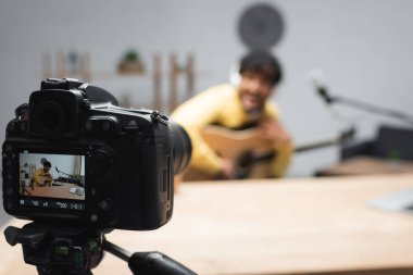 Podcast stüdyosundaki masada, mikrofonun yanında akustik gitar ve dizüstü bilgisayar tutan bulanık ve genç Hintli podcaster 'ın yanında duran tripod üzerindeki dijital kamera ekranı. 