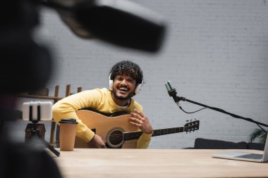 Mikrofonun yanında akustik gitar, tripodun üzerinde bulanık akıllı telefon ve stüdyoda ahşap masaya koymak için kahve tutan pozitif Hint podcaster 'ı. 