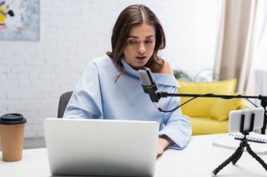 Bluz giymiş esmer yayıncı mikrofonun yanında konuşuyor ve bilgisayarını kahvenin yanında kullanıyor. Stüdyoda akış sırasında defter, kalem ve akıllı telefon kullanıyor. 