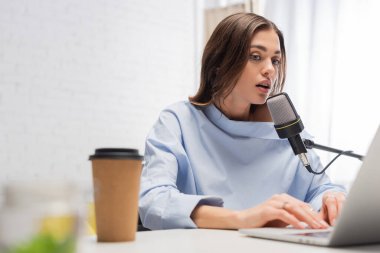 Esmer yayıncı mikrofonda konuşuyor ve kahvenin yanında bulanık dizüstü bilgisayar kullanıyor. Podcast stüdyosundaki yayın sırasında masanın üzerindeki kağıt bardağa konuyor. 