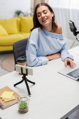 Neşeli ve bulanık podcaster mikrofonun yanındaki akıllı telefona bakıyor ve stüdyodaki akıntı sırasında laptopun yanında kahve içiyor. 